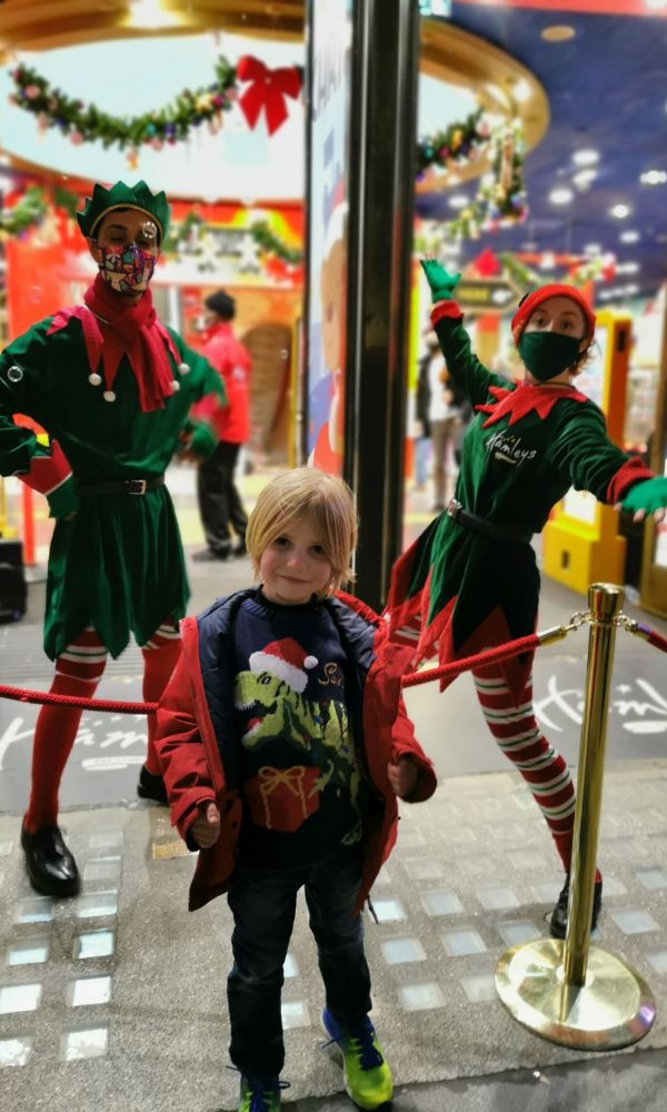Little boy in a Christmas jumper standing with the Hamleys Elves in London.