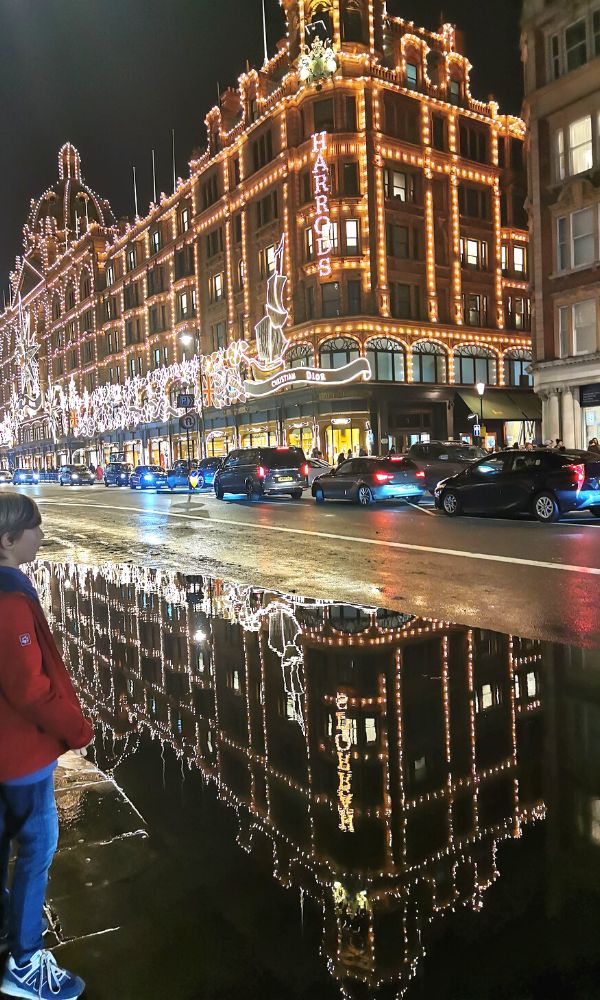 Young boy standing beside a large puddle in front of Harrods with the lights of Harrods reflecting in the puddle.