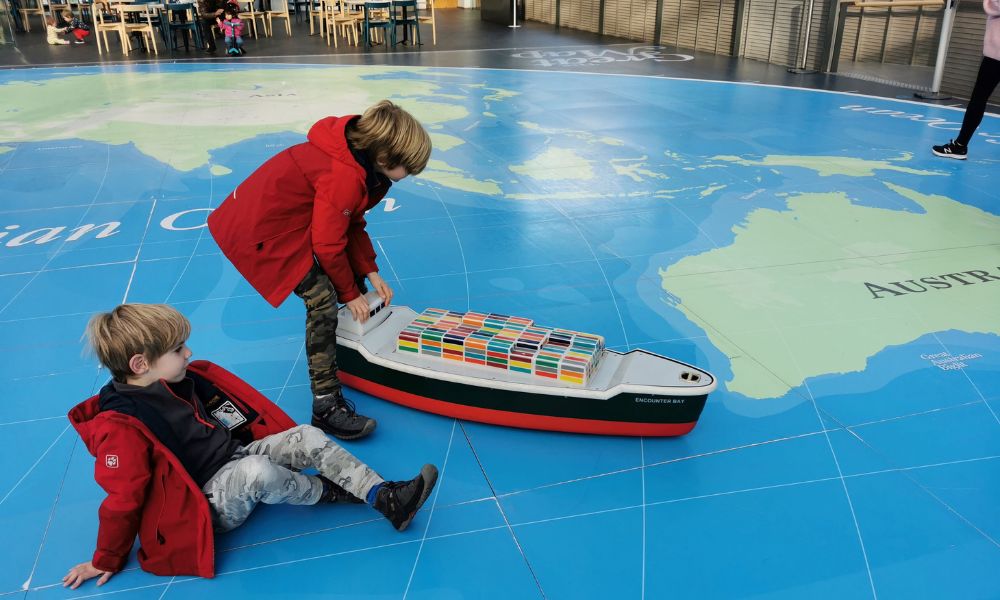 Kids in red jackets pushing a large toy cargo ship around on a giant atlas at the National Maritime Museum in London.