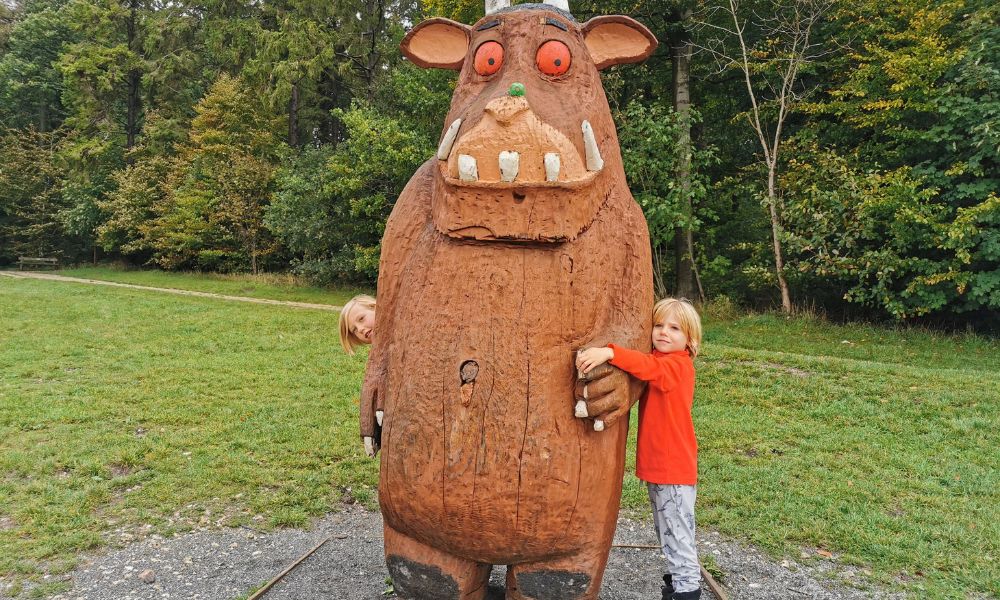 Young boy in a bright orange top hugging a large wooden gruffalo statue.